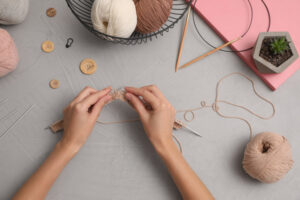 des mains en train de tricoter avec autour des boutons, du fil, des pelotes et une plante